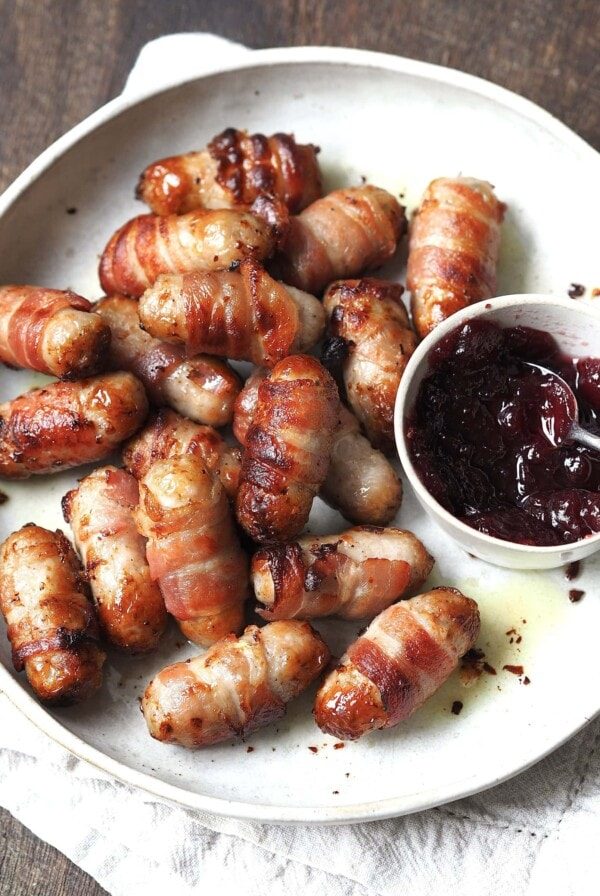 Sausages wrapped in bacon on a white serving plate next to a bowl of cranberry sauce.
