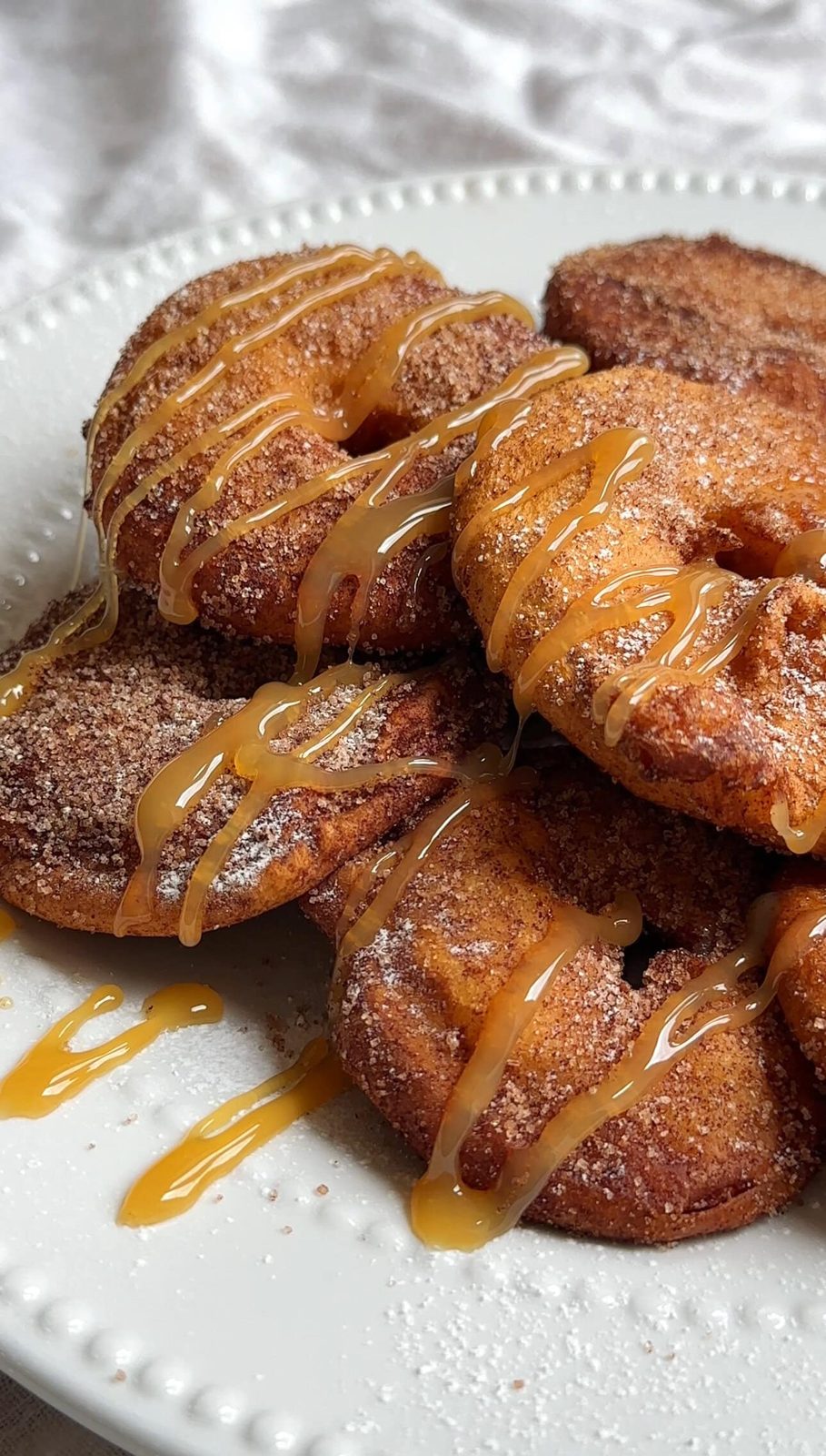Apple beignets stacked on plate with cinnamon sugar and caramel sauce drizzle.