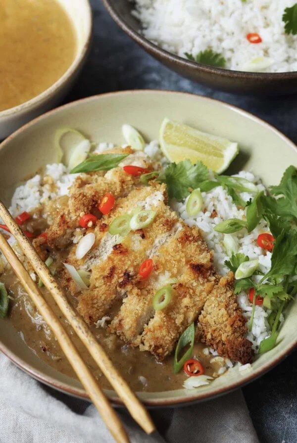 Chicken katsu cutlet in slices served over a bowl of rice with spring onions, chilli slices, lime wedges and coriander as a garnish.