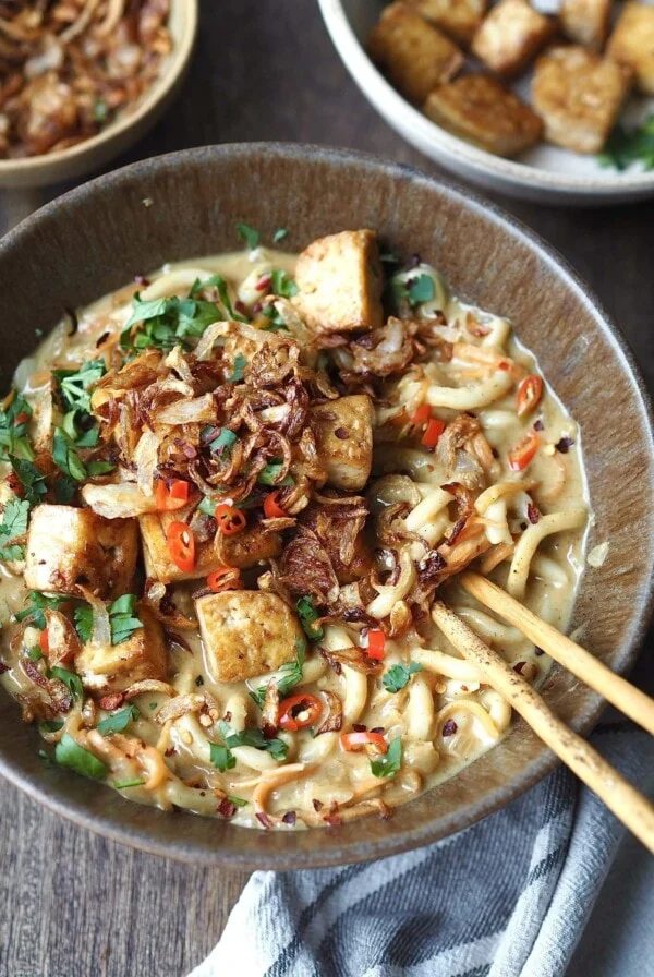 Bowl of curry udon topped with tofu and crispy shallots being eaten with chopsticks.