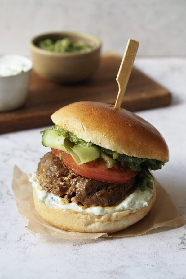 Lamb burger with tomato, cucumber ribbons, tzatziki and pesto on a white marble countertop.