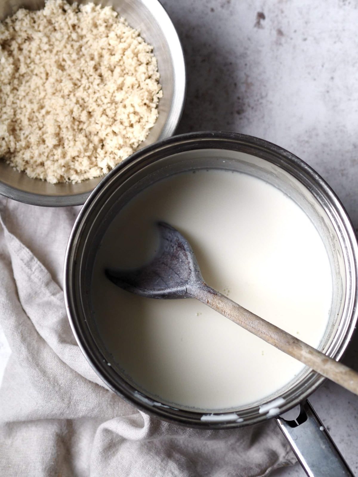 A saucepan of milk next to a bowl of breadcrumbs.