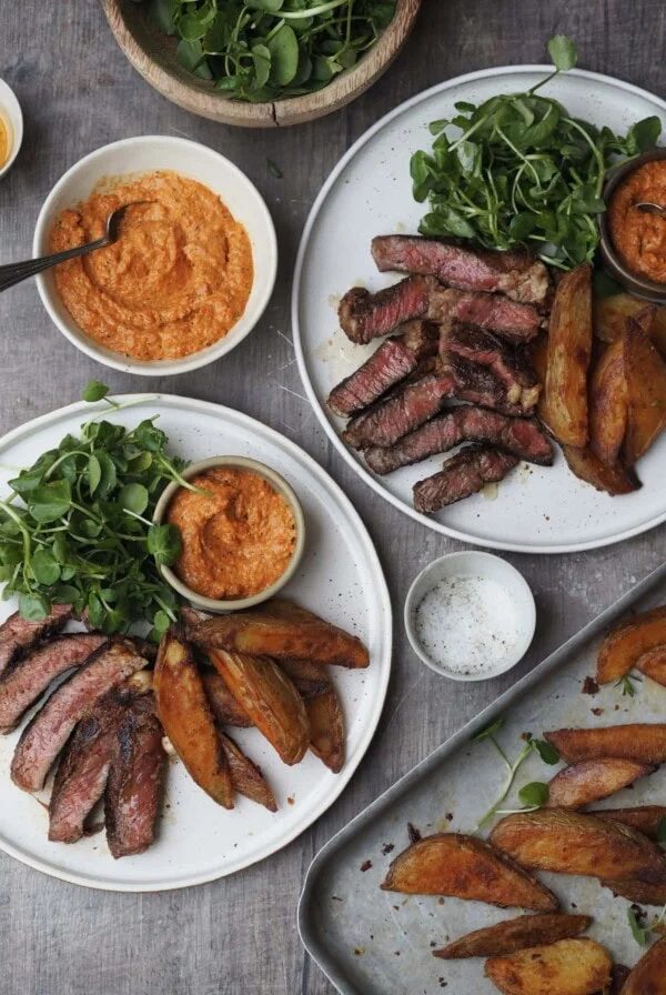 Plates of sliced rib eye steak with watercress, potato wedges and romesco sauce.