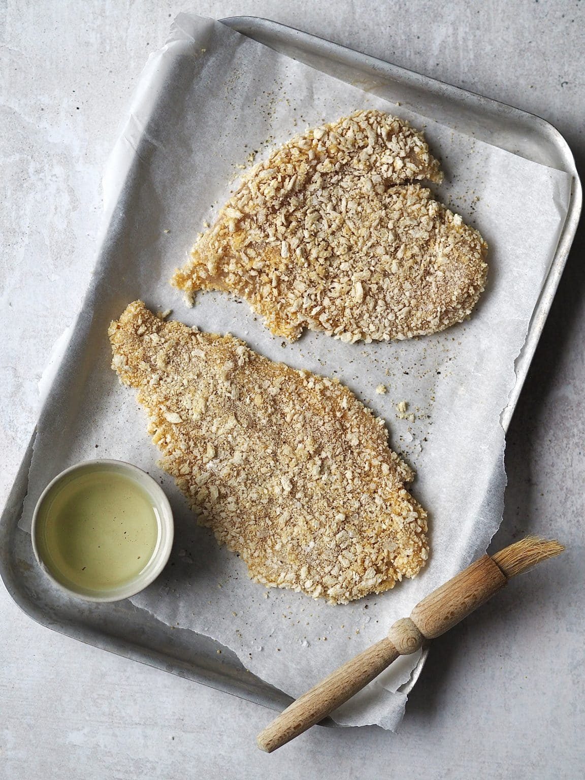 Breaded chicken cutlets on a lined baking tray being brushed with a bowl of oil.
