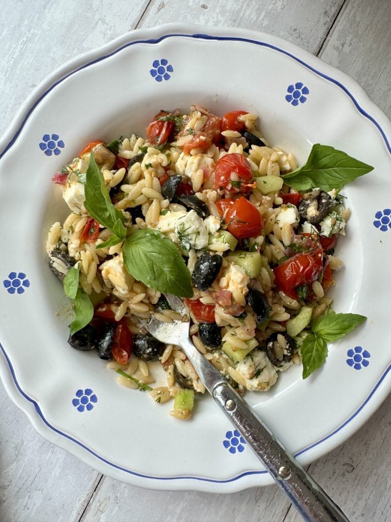 Greek pasta salad in white bowl with blue flowers with a fork in the plate.