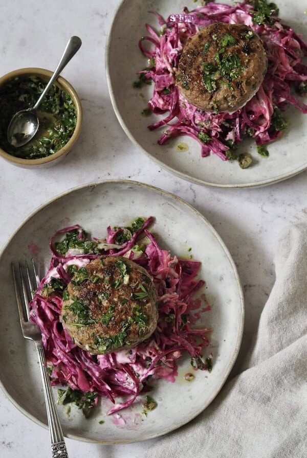 Chicken burgers on piles of pink yoghurt and beetroot slaw on grey stone plates next to a small bowl of salsa verde that's been spooned over the top.