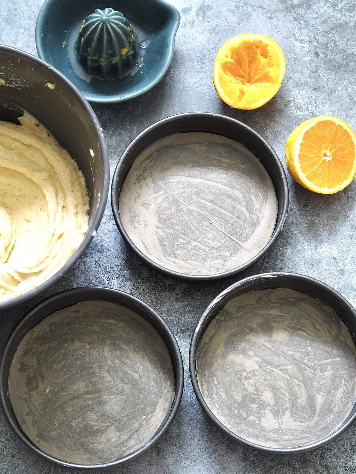 Greased round cake pans on table with cake batter and oranges.