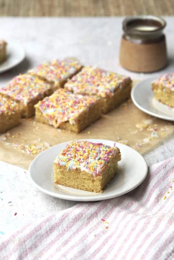 Slice of rainbow sprinkle iced sponge on a white plate with the rest of the cake in the background.