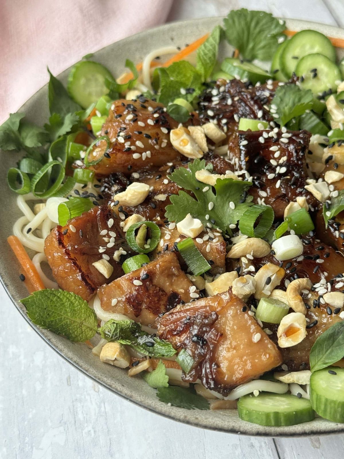 Pork belly noodles in a white bowl decorated with herbs and cucumber.