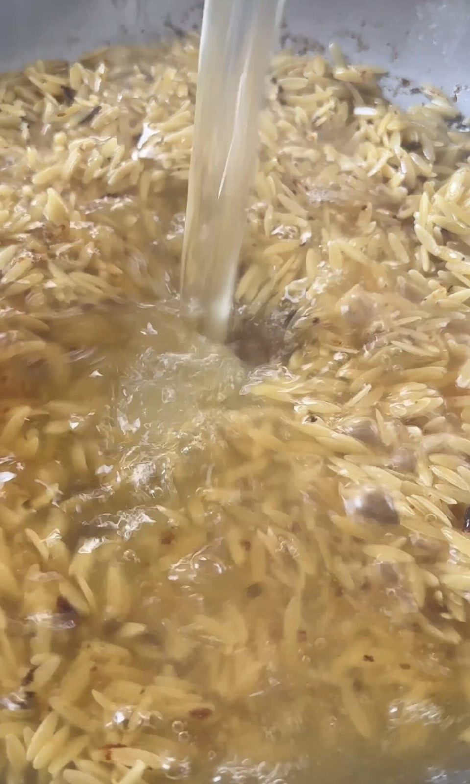 Pouring chicken stock into pan of toasted orzo.