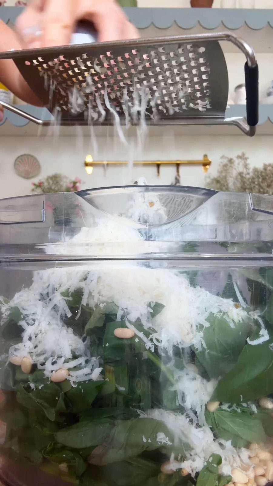 Grating Parmesan over food processor bowl with basil, pine nuts, and other pesto ingredients.
