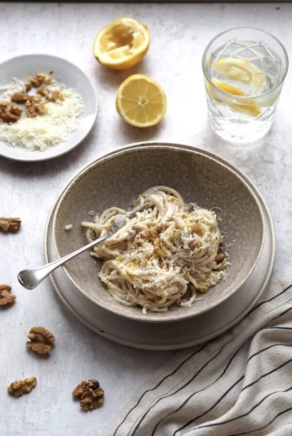 Grey bowl of spaghetti with white pesto with a glass of lemon water and a grey plate of toppings in the background.