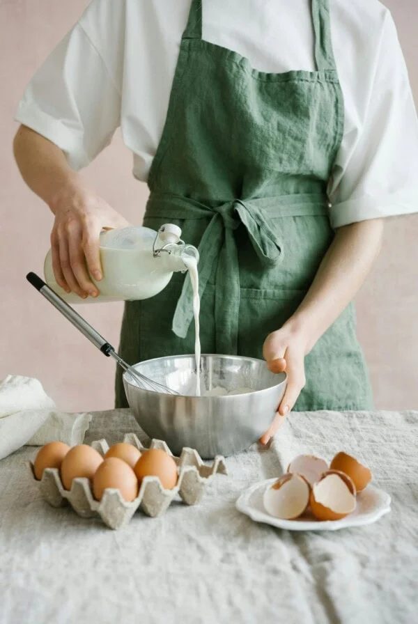 Woman pouring milk from a glass bottle into the bowl with eggs in front.