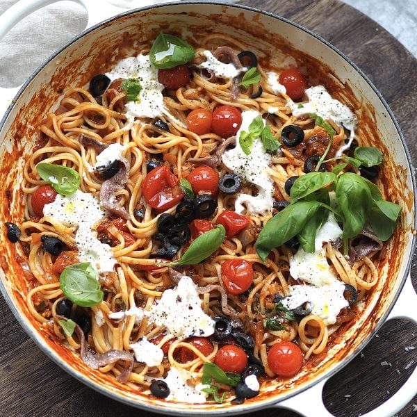 White shallow casserole with puttanesca dotted with torn burrata and fresh basil.
