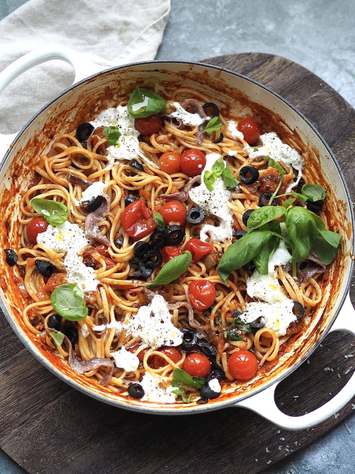 White shallow casserole with puttanesca dotted with torn burrata and fresh basil.