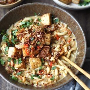 Bowl of curry udon topped with tofu and crispy shallots being eaten with chopsticks.