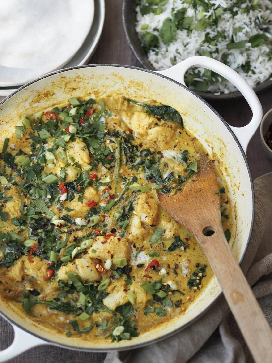 White shallow casserole dish with a fish and spinach curry next to serving bowls and a bowl of herbed rice.