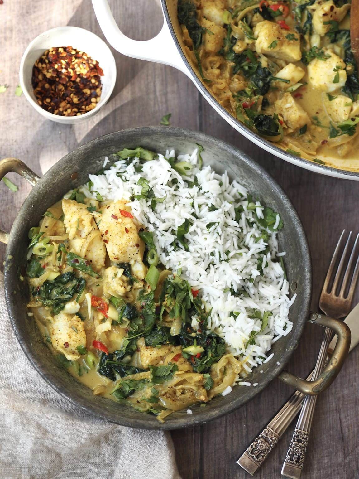 A metal serving dish of cod curry with coriander rice next to a bowl of dried chilli flakes.