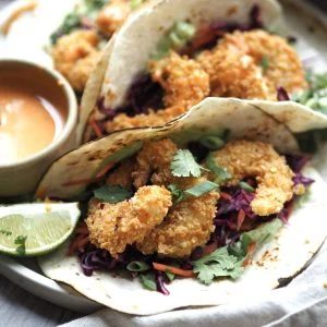 Close up of panko prawn tacos with lime wedges and a bowl of sriracha sauce on the side.