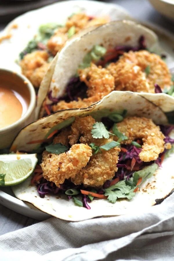 Close up of panko prawn tacos with lime wedges and a bowl of sriracha sauce on the side.