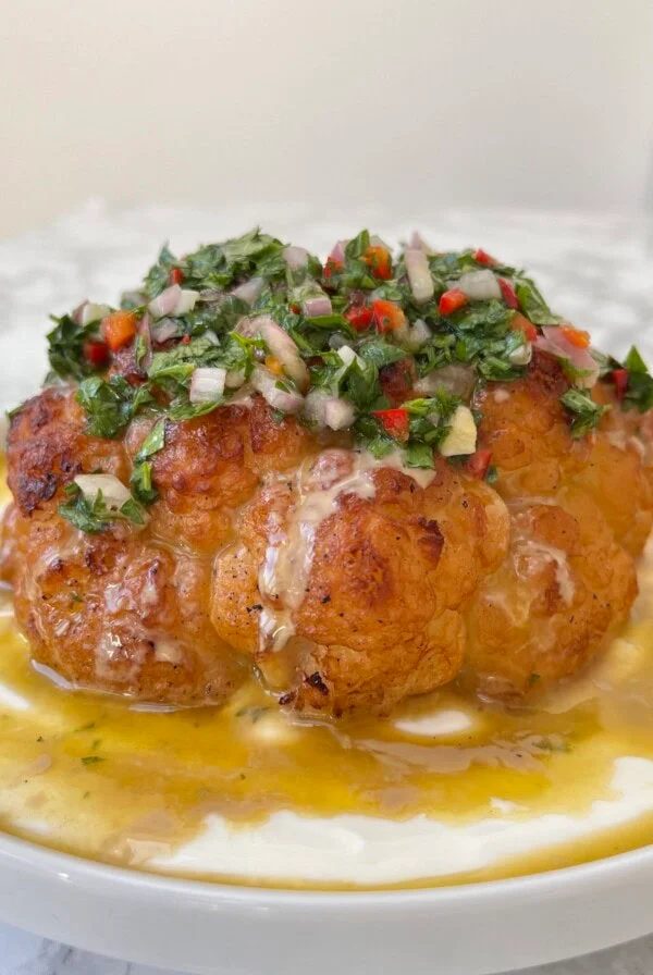 Close up of a whole roasted cauliflower with tahini sauce and chimichurri spooned over the top.