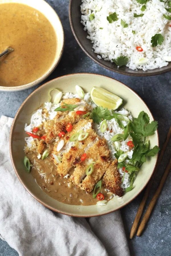 A chicken katsu curry bowl next to serving bowls of herbed rice and Japanese coconut curry sauce.