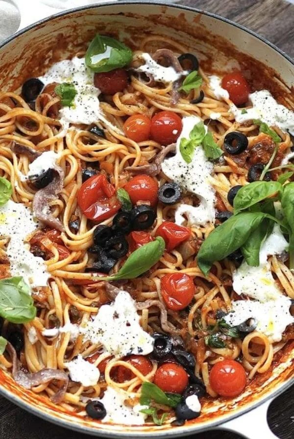 White shallow casserole with puttanesca dotted with torn burrata and fresh basil.