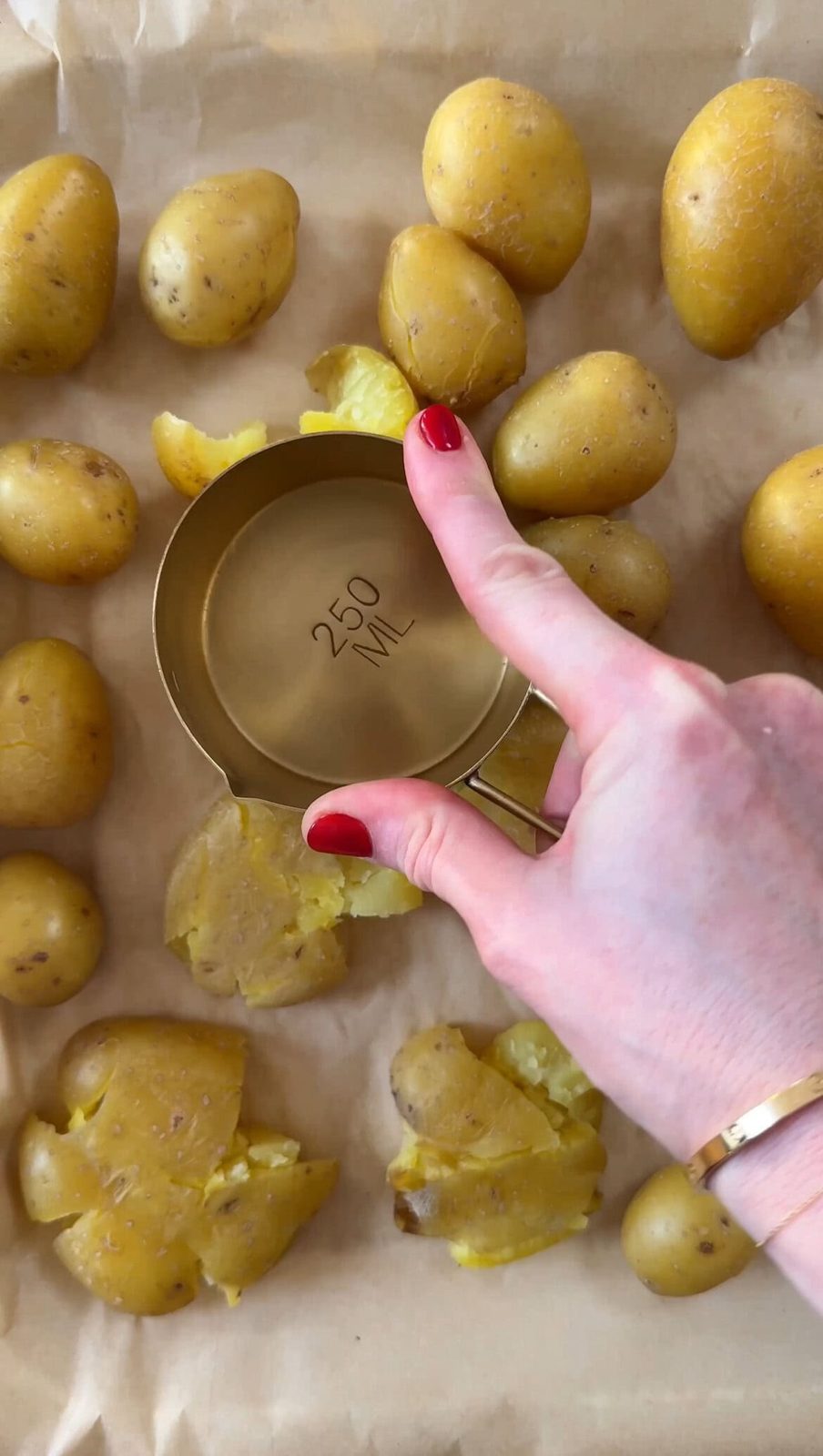 Smashing boiled baby potatoes with measuring cup on parchment-lined baking sheet.