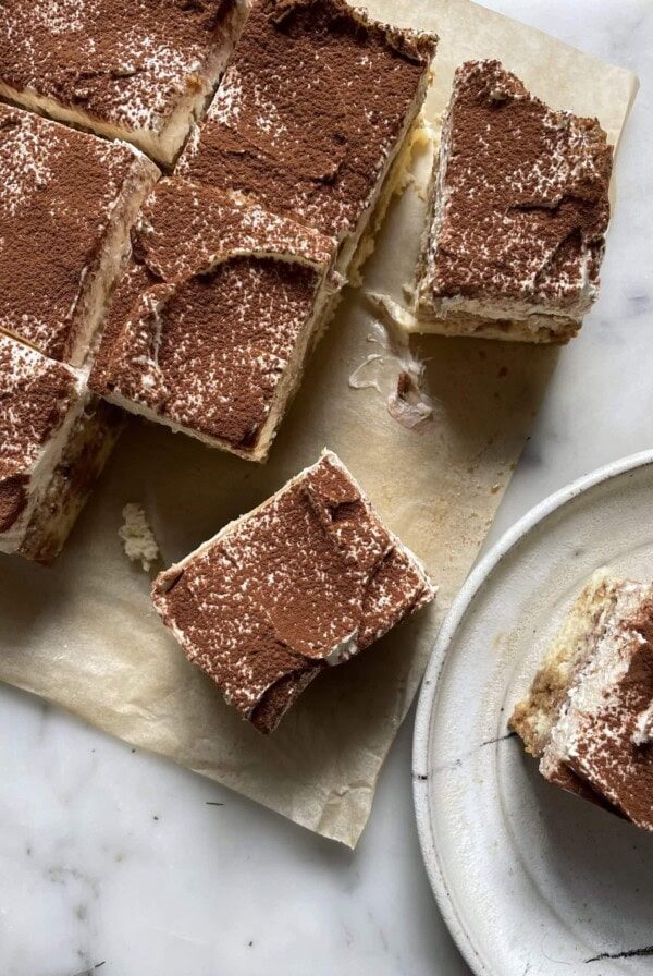 A slice of tiramisu tres leches cake on a plate with the full cake on the table