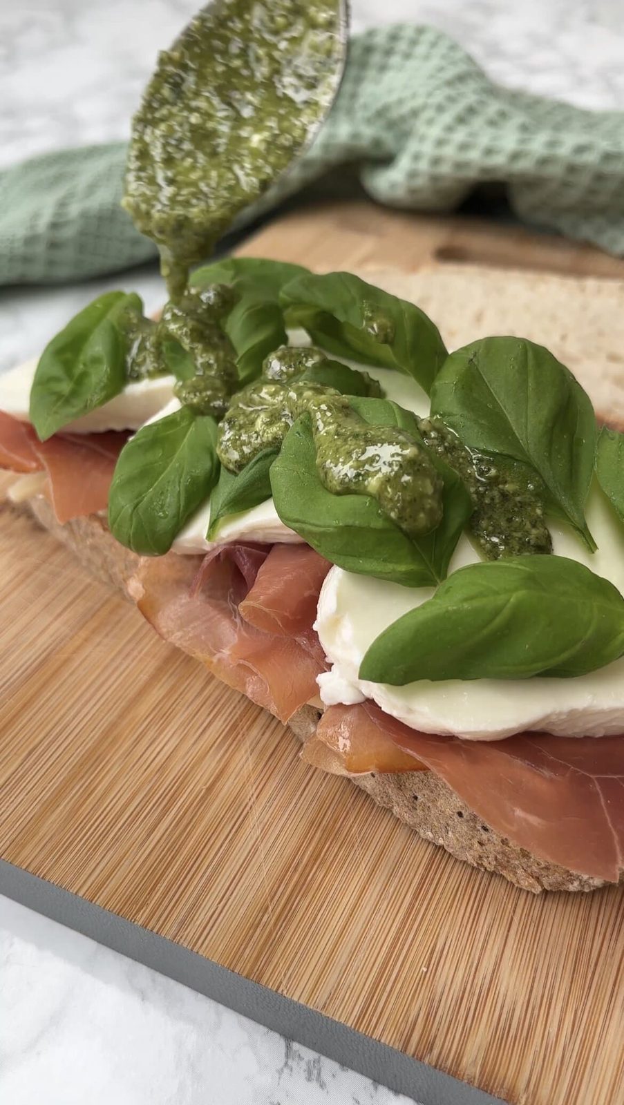 Pouring pesto over sandwich with meat, cheese, and fresh basil.