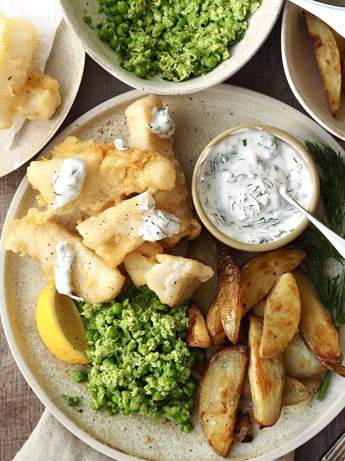 Fish goujons on a plate with potato wedges, lemon and peas drizzled with homemade tartare sauce.