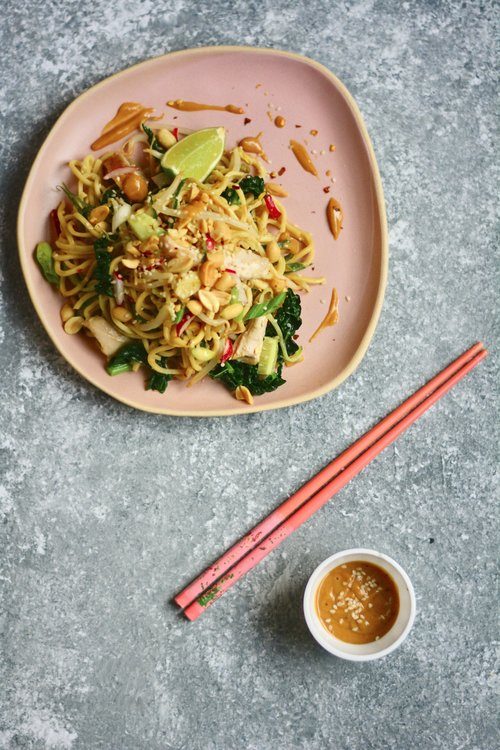 Plate with Thai-inspired peanut noodles and chicken near chopsticks and bowl of peanut sauce.