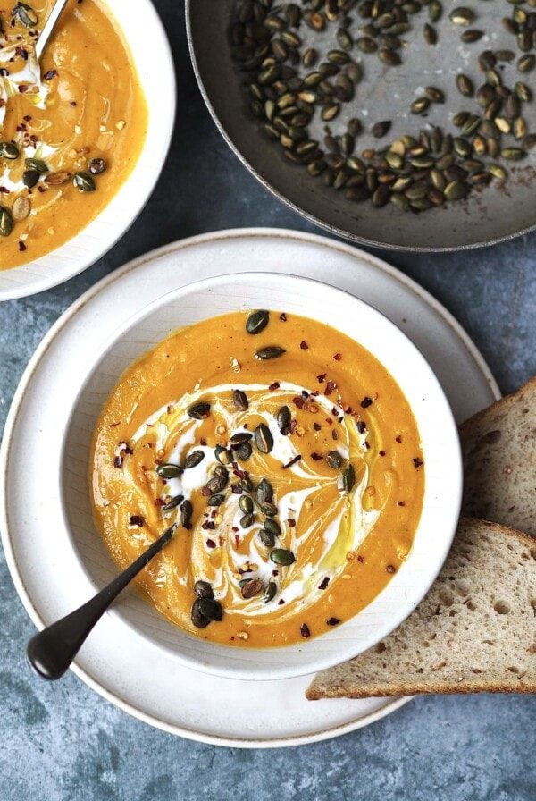 White bowl of butternut squash soup garnished with pumpkin seeds and chilli flakes with bread on the side.