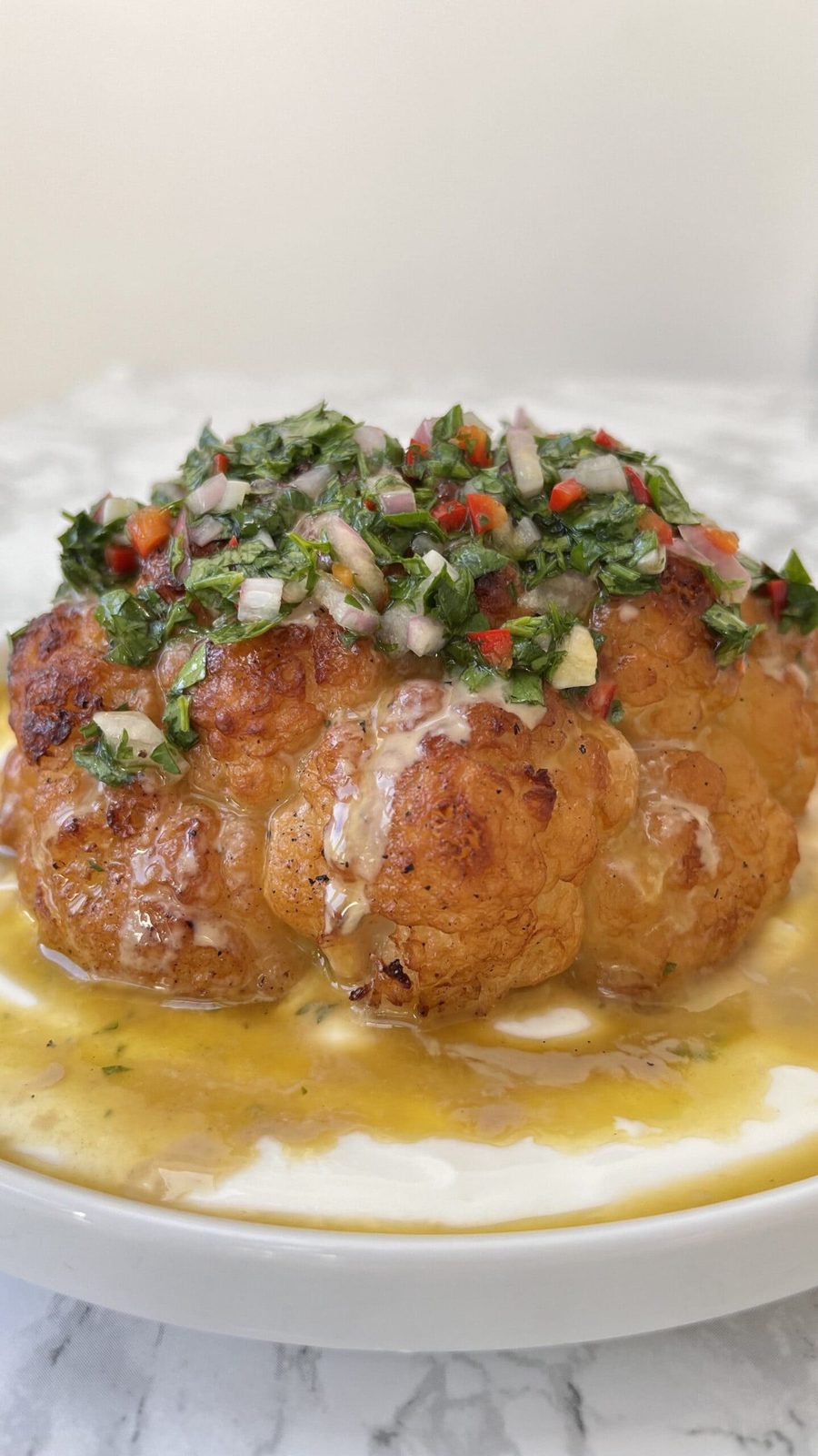 Close up of a whole roasted cauliflower with tahini sauce and chimichurri spooned over the top..