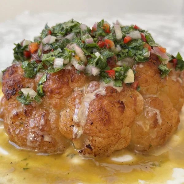 Close up of a whole roasted cauliflower with tahini sauce and chimichurri spooned over the top.