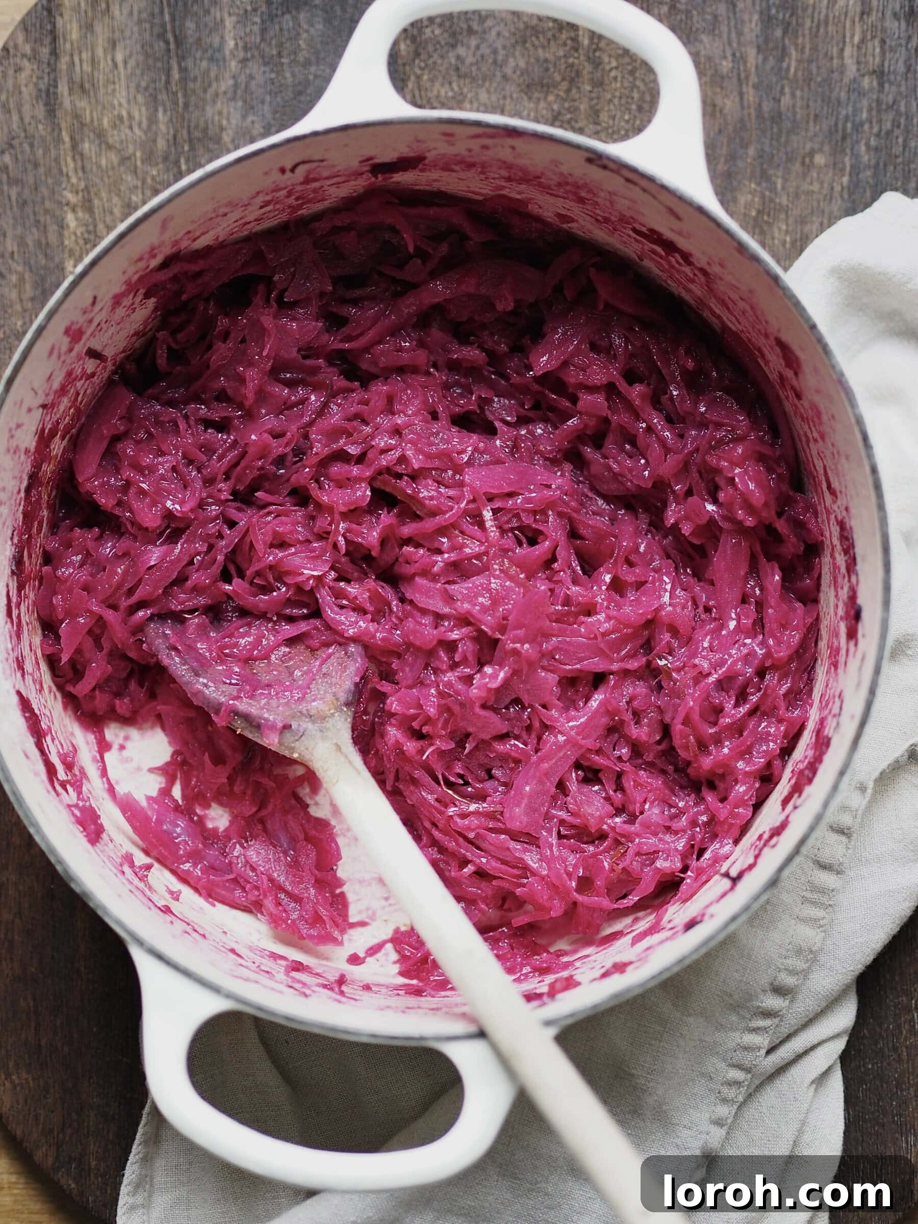 Cooked red cabbage and apple mixture being stirred with a wooden spoon in a white casserole dish, showing its tender texture.