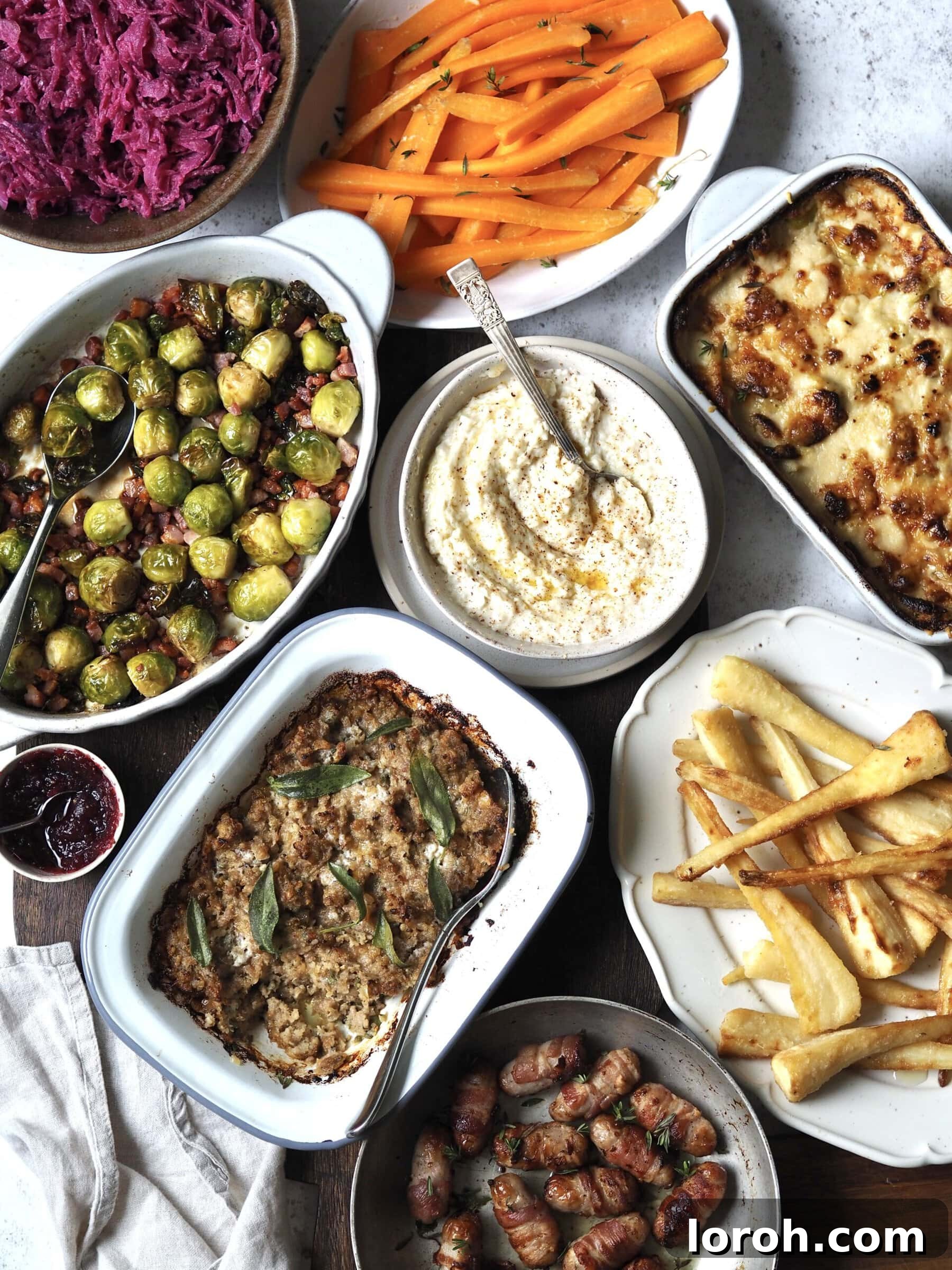 An array of delicious Christmas side dishes in serving bowls, including red cabbage, maple-glazed carrots, Brussels sprouts with bacon, bread sauce, cheesy leeks, baked stuffing, pigs in blankets, and roasted parsnips.