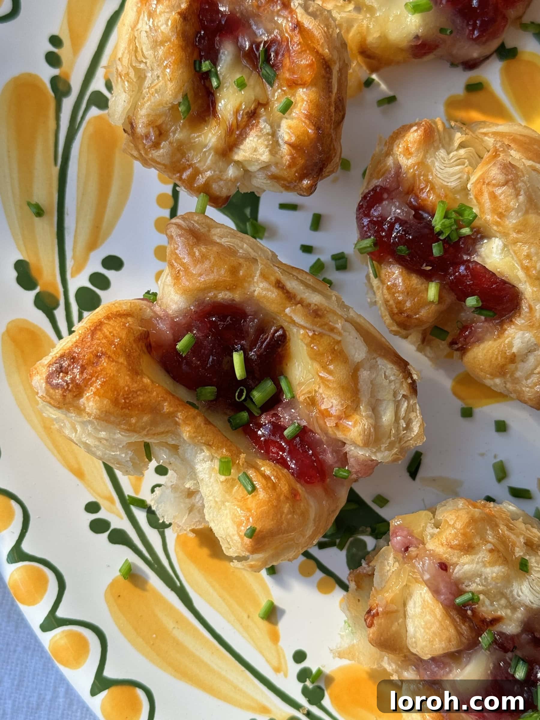 Golden Flaky Brie Bites 3 Close-up of baked Brie Puff Pastry Bites with melted brie and cranberry sauce, garnished with chives.