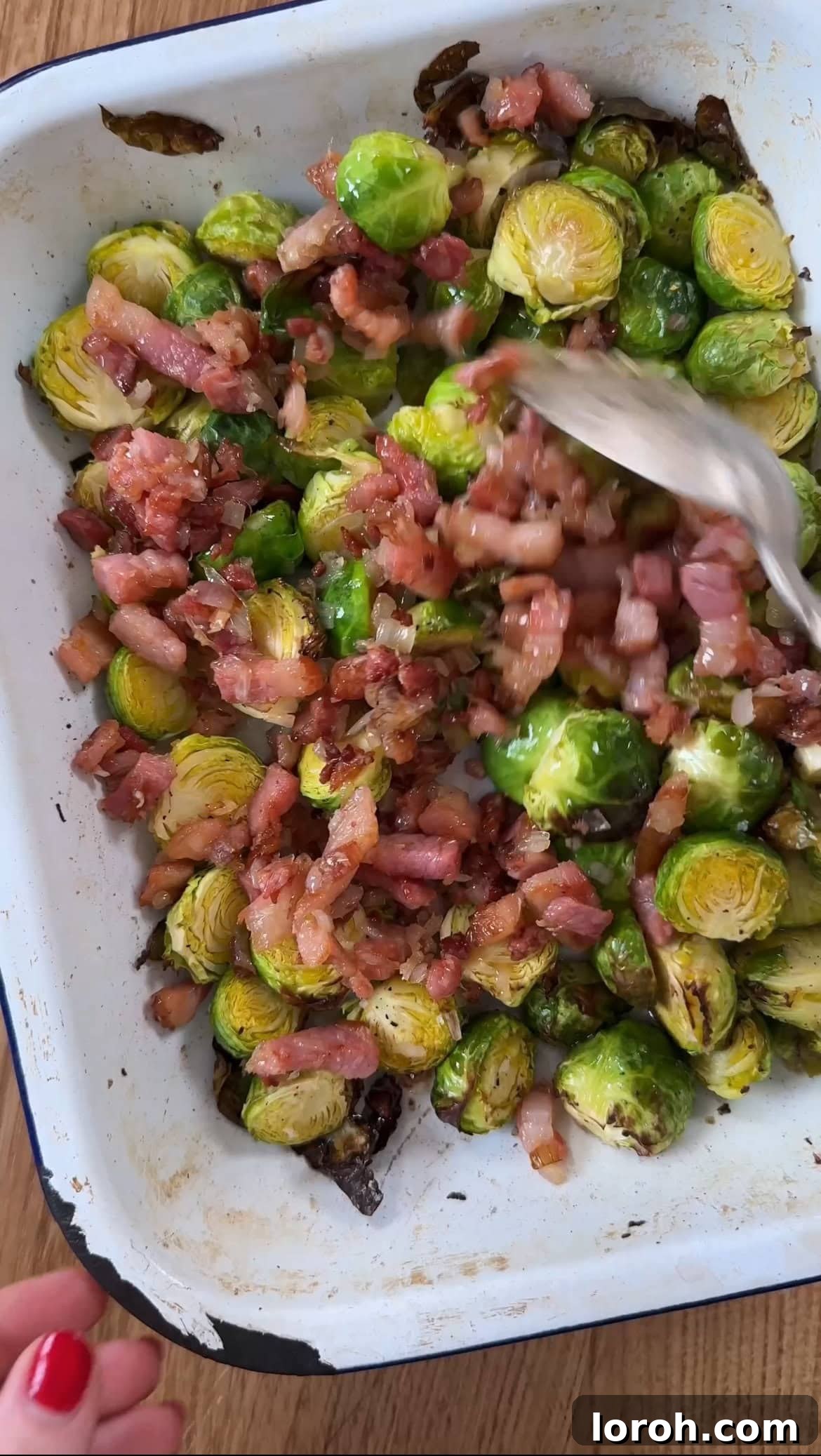 The crispy pancetta, sautéed shallot, and garlic mixture being added to the roasted Brussels sprouts in the baking dish.