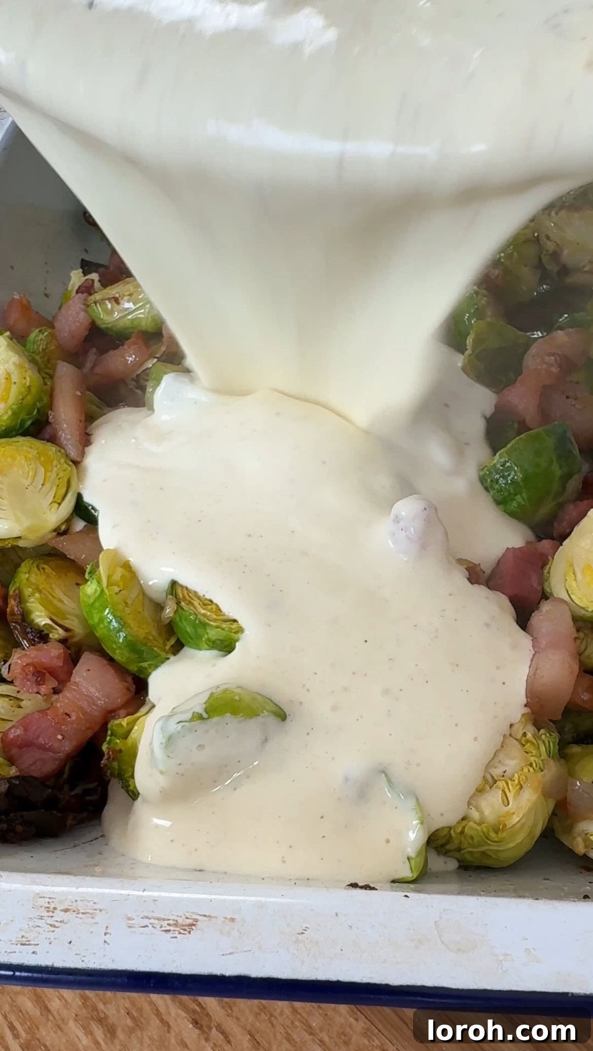 The creamy cheese sauce being poured generously over the roasted Brussels sprouts and pancetta mixture in the baking dish.