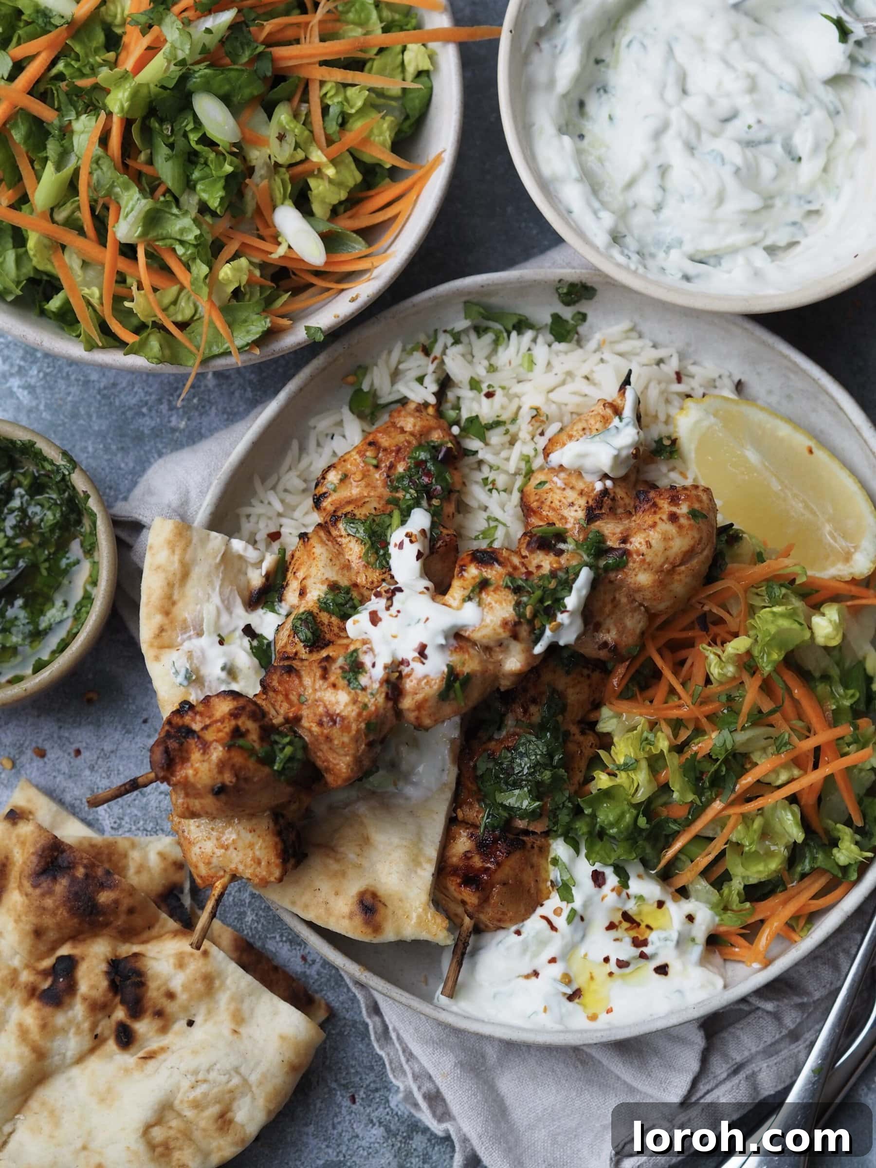 An overhead shot of baked chicken skewers, salad, rice, and raita on a rustic wooden table.