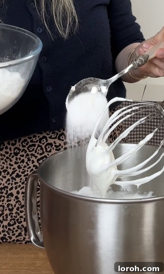 Adding caster sugar to the whipped egg whites one spoonful at a time for stabilization.