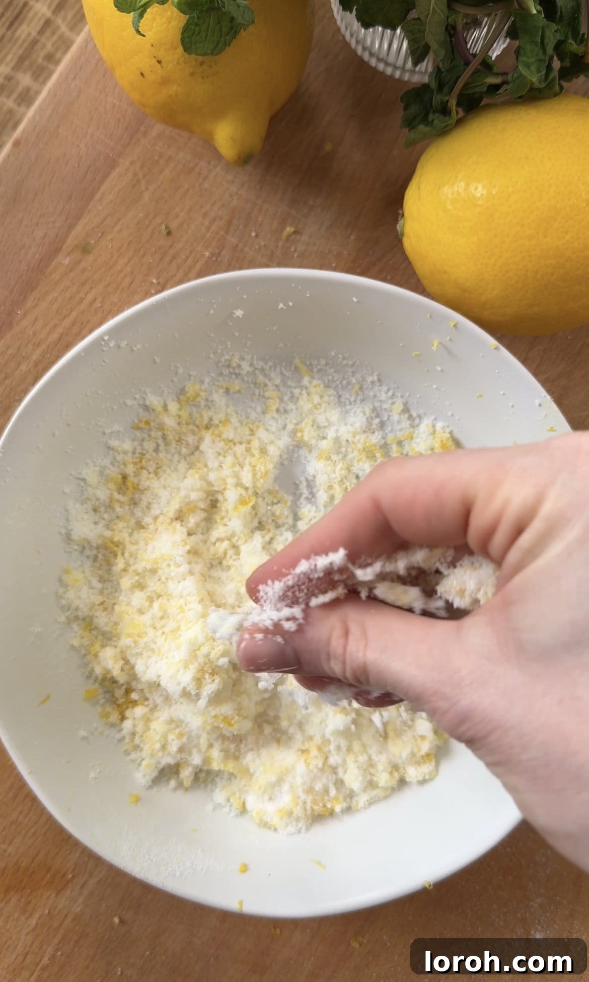 Mixing caster sugar and finely grated lemon zest together with fingertips in a glass bowl to release essential oils.