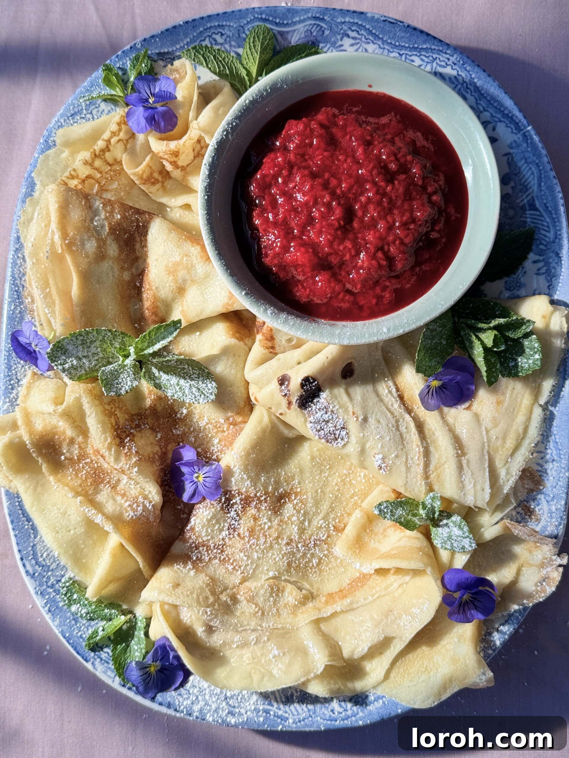 Golden Pannekaker on a blue plate, garnished with vibrant raspberry compote and a dusting of powdered sugar.