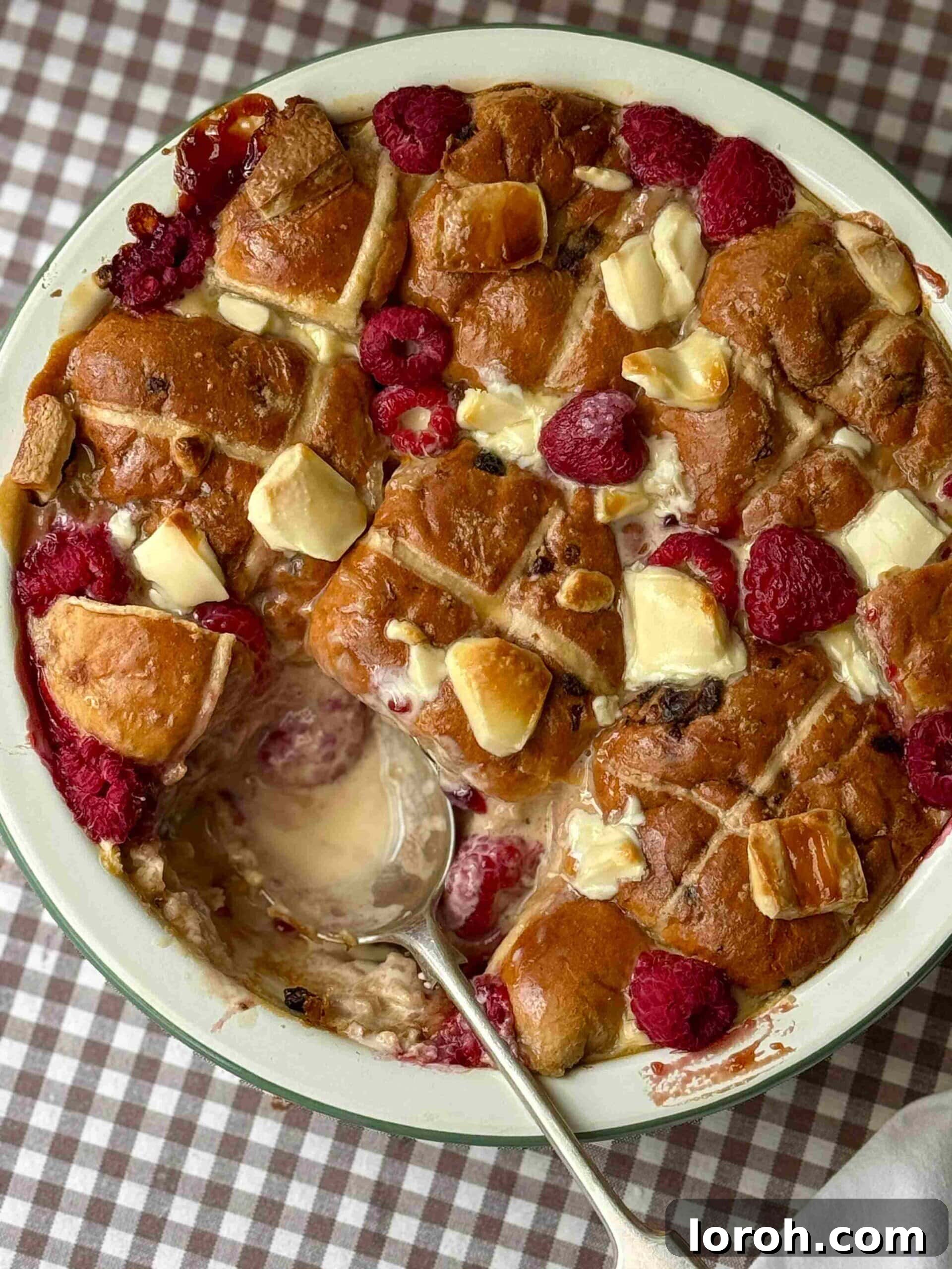 A serving of Hot Cross Bun Bread and Butter Pudding with a spoon, highlighting the soft buns, melted white chocolate, and raspberries.