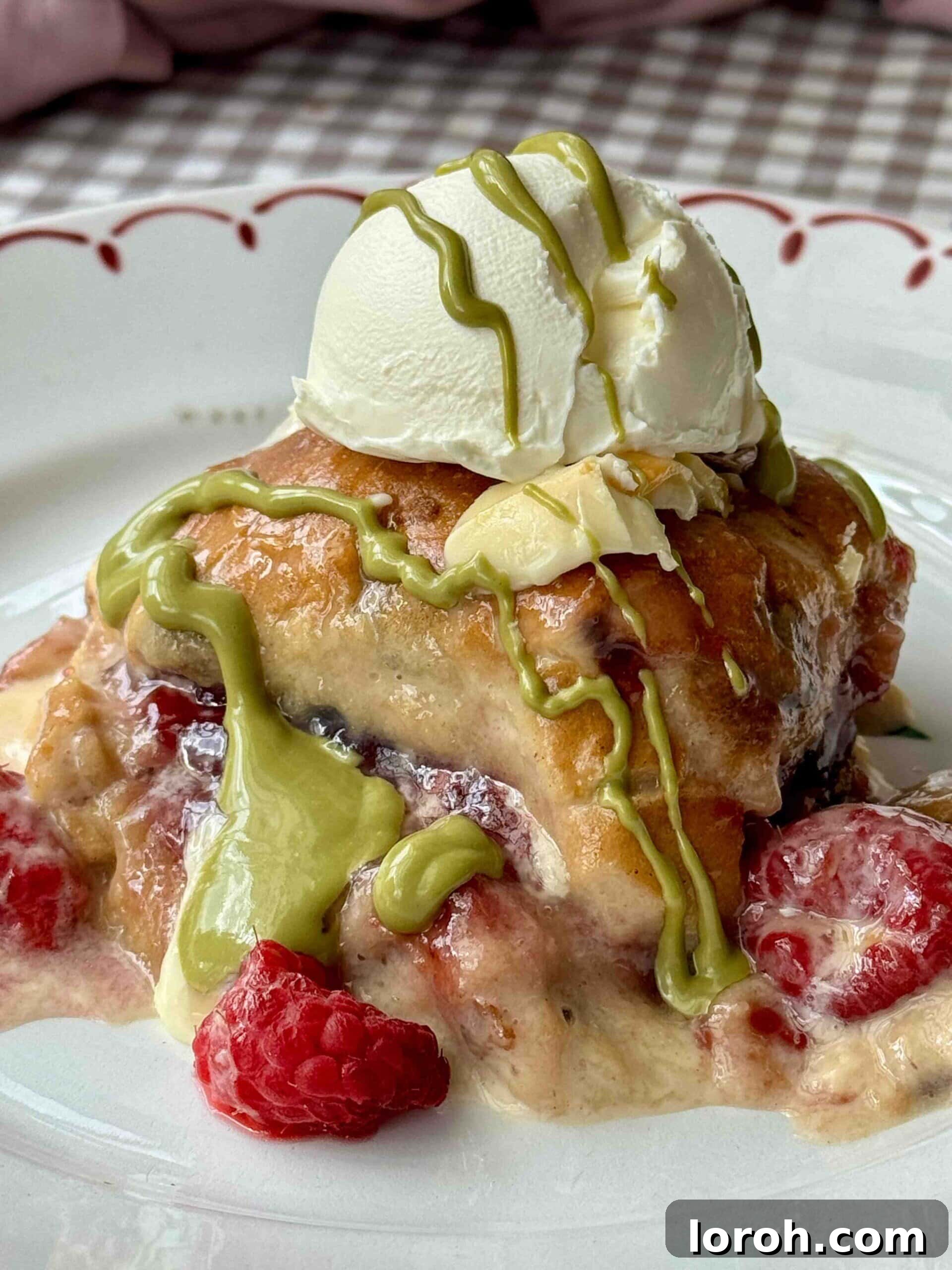 Baked Hot Cross Bun Bread and Butter Pudding adorned with white chocolate, raspberries, and a elegant drizzle of pistachio cream.