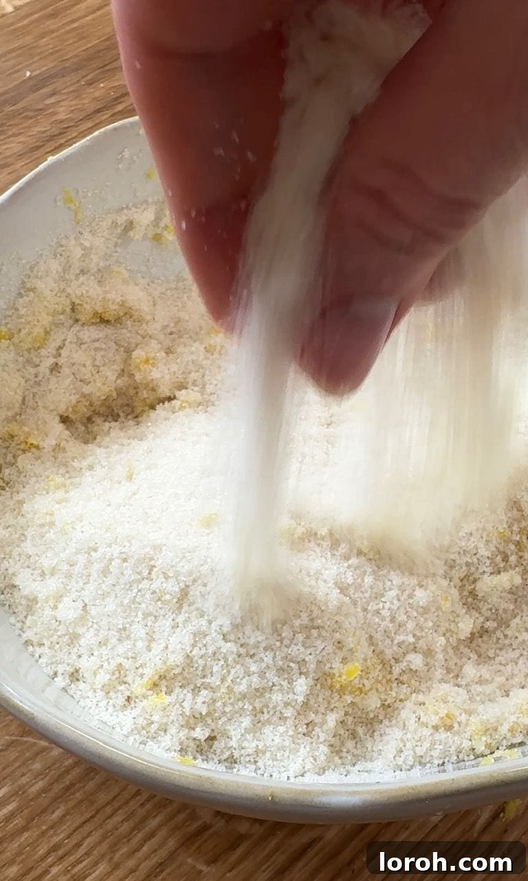 Close-up photo of lemon zest being rubbed into caster sugar in a mixing bowl, creating a fragrant mixture.