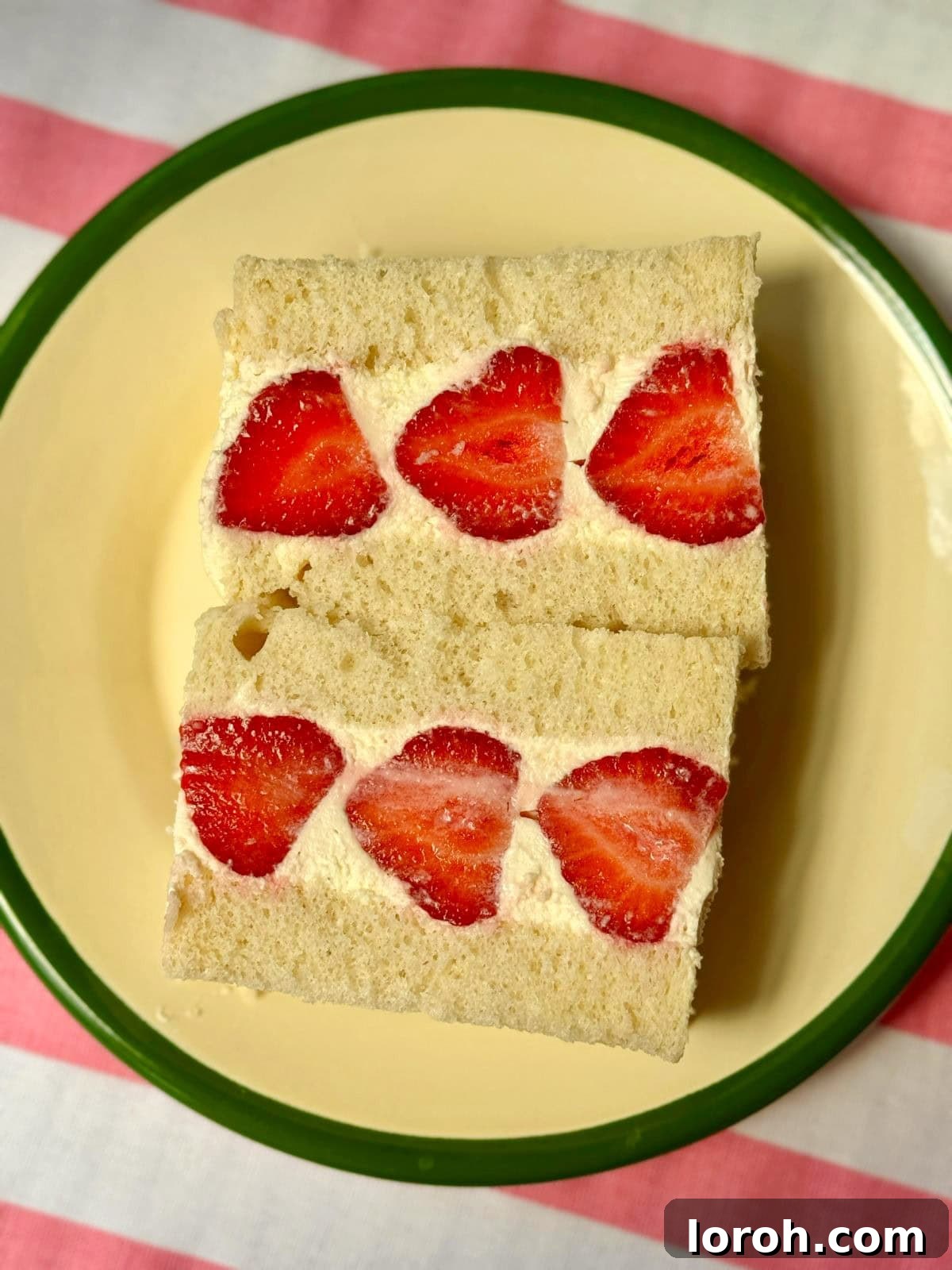 Close-up of a Japanese strawberry sandwich (Ichigo Sando) with a perfect cross-section, showing layered strawberries and cream, on a white plate with a green rim.