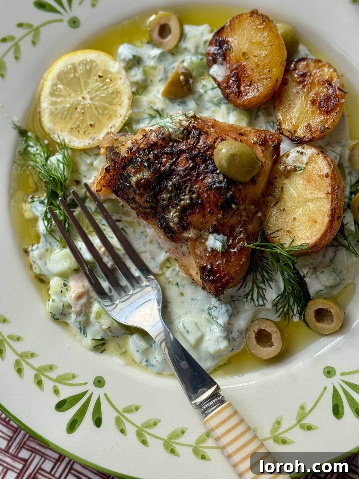 A serving of Greek chicken and lemon potatoes on a white plate, garnished with fresh herbs and a dollop of cucumber yogurt.