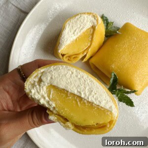 A vibrant mango pancake, delicately thin and yellow, held up to showcase its sweet mango and cream filling.